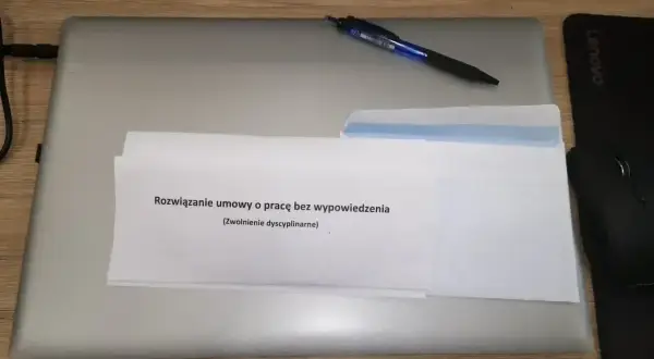 Na biurku leży laptop, na nim dokument z napisem "Rozwiązanie umowy o pracę bez wypowiedzenia (zwolnienie dyscyplinarne)". Obok długopis i myszka.