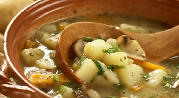 Zupa ziemniaczana z grzybami i marchewką w glinianej misce, nabierana drewnianą łyżką.