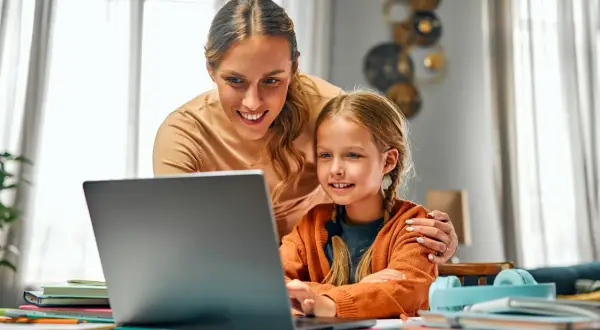Uśmiechnięta mama pomaga córce w nauce przy laptopie. To przykład troskliwej opieki na dziecko 2 dni w tygodniu, gdy rodzice pracują.