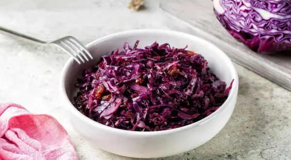 Un bol de chou rouge recette, garni de raisins secs, prêt à être dégusté. Une moitié de chou rouge est visible sur une planche à découper en arrière-plan.