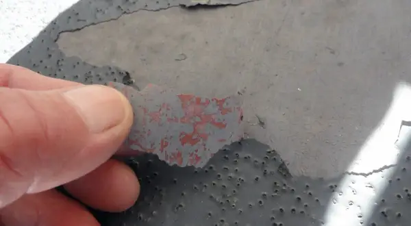 Dedo retirando capa desprendida de pintura gris sobre hormigón con picaduras, indicando necesidad de reparación de hormigón.