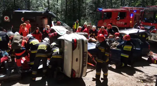 Akcja ratunkowa po wypadku. Strażacy KSRG zabezpieczają teren i wyciągają poszkodowanych z przewróconego auta. W tle wozy strażackie i karetka.