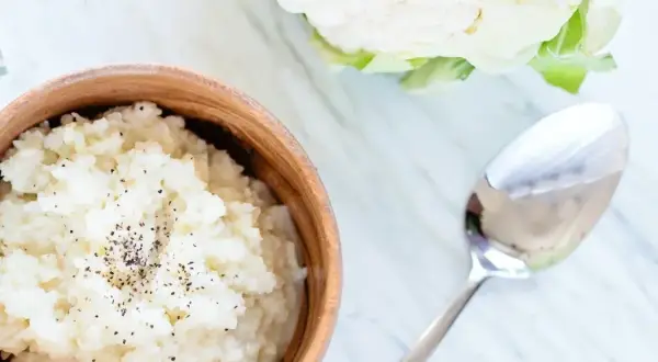 Pyszne puree z kalafiora w drewnianej misce, posypane pieprzem. Obok leży łyżka, a w tle świeży kalafior.