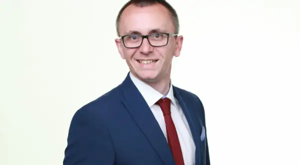 Mężczyzna w garniturze i okularach, uśmiecha się na białym tle.