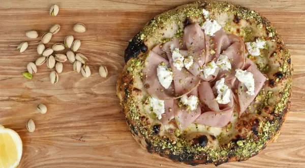Pizza mit Mortadella, Pistazien und cremigem Käse, daneben einige Pistazien und eine Zitronenspalte.