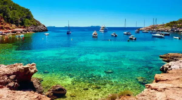 Aguas cristalinas en cala salinas, con barcos anclados y una costa rocosa y verde.