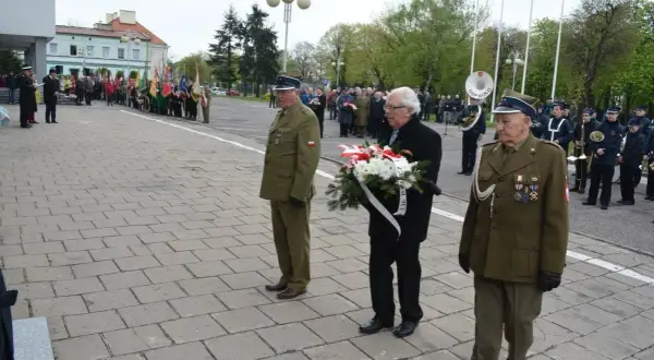 Trzech mężczyzn w mundurach wojskowych i cywilnych, jeden z nich niesie wieniec. W tle parada i orkiestra.