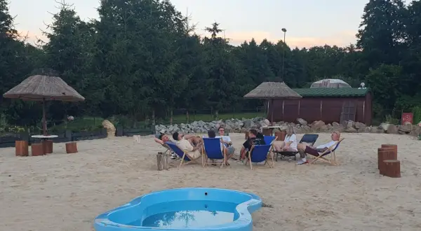 Relaks na piasku w Krężnicy Jarej. Grupa przyjaciół odpoczywa na leżakach przy małym basenie, w tle drewniana sauna i palmy.