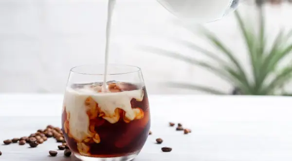 Mjölk hälls i ett glas med iskaffe, perfekt för ett enkelt iskaffe recept. Kaffebönor ligger utspridda bredvid.
