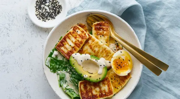 Sałatka z grillowanym serem halloumi, awokado, jajkiem i zielonymi liśćmi, posypana sezamem.