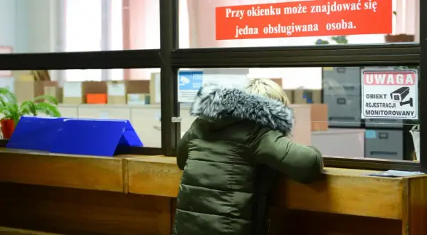 Osoba w zielonej kurtce przy okienku urzędu, znak "Uwaga obiekt monitorowany".