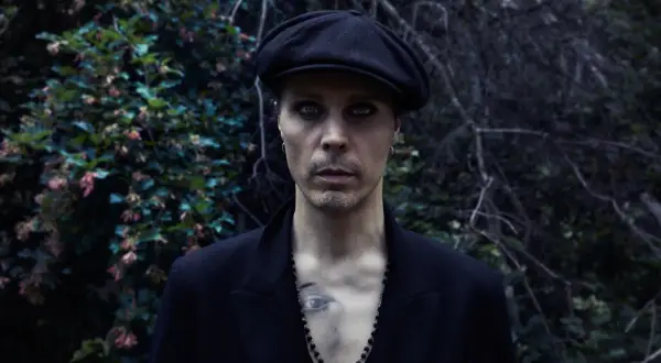 Ville Valo w czarnym kaszkiecie i marynarce, z intensywnym makijażem oczu, stoi na tle ciemnej, leśnej roślinności.