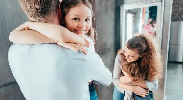 Dziewczynka mocno przytula tatę, jej uśmiech świadczy o dobrym samopoczuciu i zdrowej psychice. Mama tuli synka w tle.