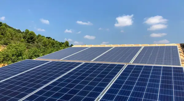 Mi experiencia con placas solares: paneles azules captan la luz del sol en un tejado, con colinas verdes y cielo azul de fondo.
