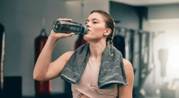 Eine Frau trinkt nach dem Training aus ihrer Trinkflasche. Sie hat ein Handtuch um den Hals.