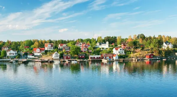 Vackra röda och vita hus längs en grönskande kustlinje, speglande sig i det blå vattnet. En idyllisk vy av sommar i Stockholm.