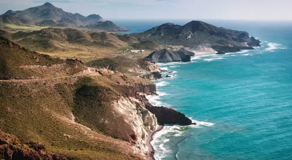 Costa escarpada cerca de Níjar, con el mar turquesa rompiendo en acantilados blancos y rojizos. Montañas verdes y una carretera serpentean en la distancia.