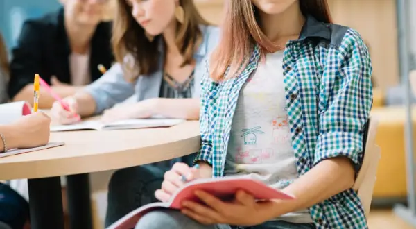Uczniowie przy stole, skupieni na nauce biologii. Jedna z dziewczyn notuje, druga czyta, trzecia uśmiecha się, trzymając zeszyt.