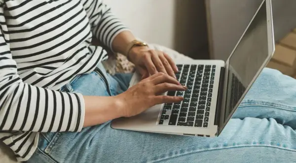 Kobieta w pasiastym swetrze i jeansach pracuje na laptopie, co symbolizuje nienormowany czas pracy i elastyczność.