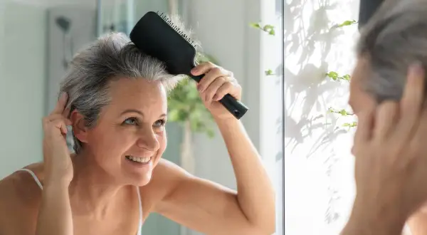 Mujer sonriente se peina el cabello gris en el espejo, lista para matizar canas y lucir radiante.
