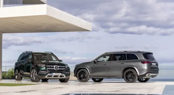 Dwa Mercedesy GLS 2025, jeden zielony, drugi szary, zaparkowane przed nowoczesnym budynkiem z basenem.