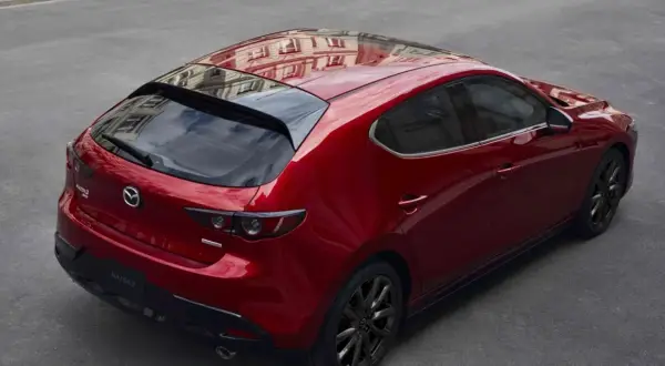 Czerwony hatchback Mazda 3 z czarnymi felgami, odbijający budynek na dachu. Wygląda elegancko, w przeciwieństwie do nudnego sedana.