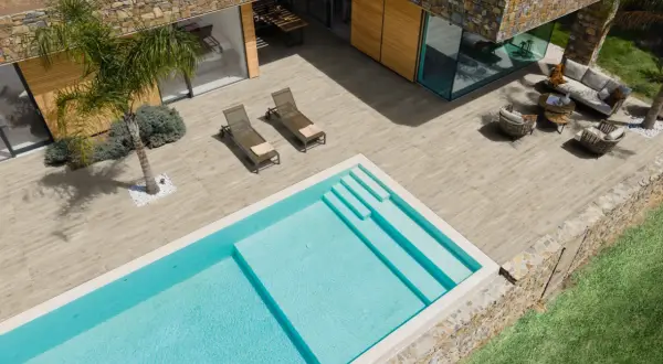 Una piscina azzurra e una moderna pavimentazione esterna in legno creano un'oasi di relax con arredi da esterno e una palma.