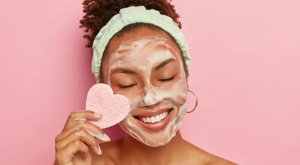 Mujer sonríe con espuma en la cara, usando una esponja en forma de corazón para su limpieza facial.