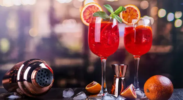Dwa orzeźwiające drinki z prosecco, ozdobione plasterkami pomarańczy i miętą, stoją obok shakera i owoców.
