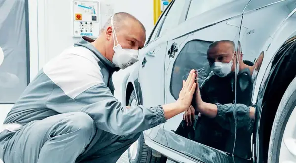 Un operaio con mascherina controlla la carrozzeria lucida di un'auto, pronto per carteggiare auto e rifinire la verniciatura.