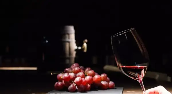 Czerwone winogrona i kieliszek wina, a w tle beczka. Czy wino bezalkoholowe ma alkohol? To pytanie nurtuje wielu miłośników tego trunku.