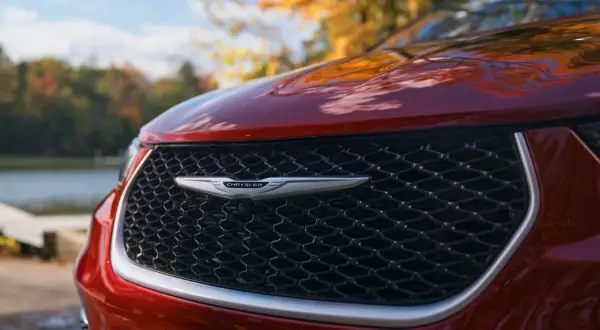 En röd Chrysler-bil med sitt ikoniska märke framtill, parkerad vid en sjö under höstens färger.
