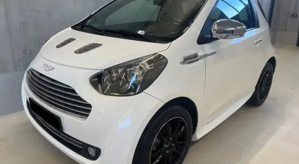 Biały Aston Martin Cygnet z czarnymi felgami i chromowanymi detalami, prezentujący się elegancko w garażu.