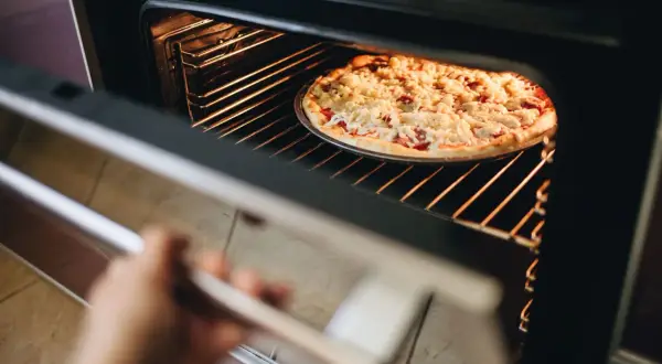 Ręka otwiera piekarnik, w którym trwa pieczenie pizzy. Gorące wnętrze rozświetla apetyczną potrawę.