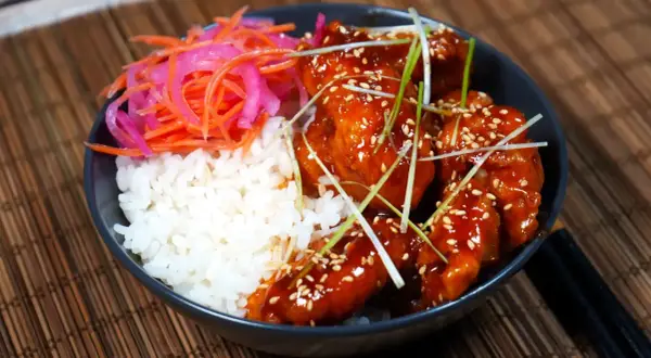 En skål med ris, krispigt koreanskt friterad kyckling i en söt och kryddig sås, toppad med sesamfrön och salladslök. Serveras med en sida av picklad morot och rödlök.