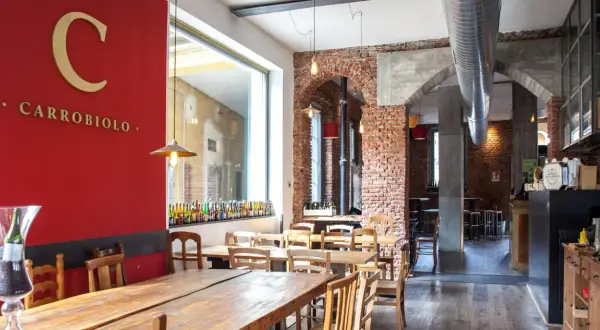 Interno di un locale con tavoli in legno e mattoni a vista. Sulla parete rossa, il logo "C CARROBIOLO" e una vetrina con tante bottiglie di birra.