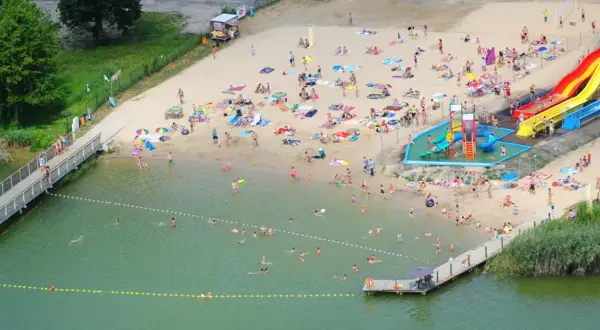 Wakacyjny dzień nad jeziorem margonińskim. Ludzie opalają się na plaży, kąpią w wodzie i korzystają ze zjeżdżalni wodnych.
