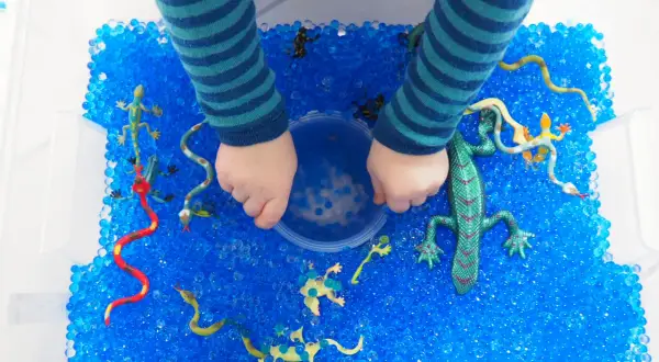 Kinderhände erkunden eine Kiste voller blauer Wasserperlen mit Spielzeug-Echsen und Schlangen. Tolle Wasserperlen Ideen für sensorisches Spiel!