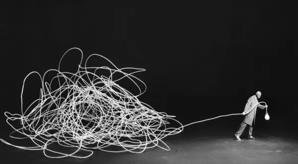 Człowiek z żarówką, jak w pracach Gilbert Garcin, ciągnie za sobą plątaninę białego sznurka na czarnym tle.