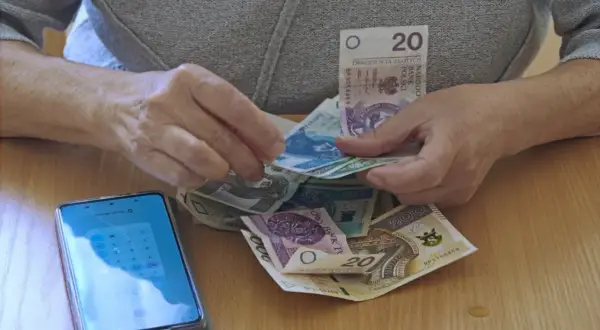 Dłonie liczą pieniądze, obok leży smartfon z kalkulatorem. Czy to dodatek pielęgnacyjny z ZUS, komu przysługuje?