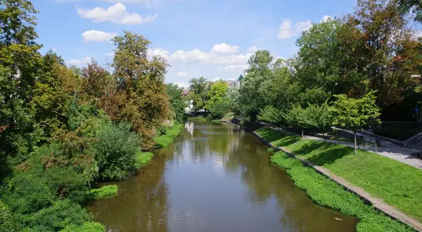 Spokojna rzeka Młynówka w Opolu, otoczona zielenią i drzewami, pod błękitnym niebem z chmurami.