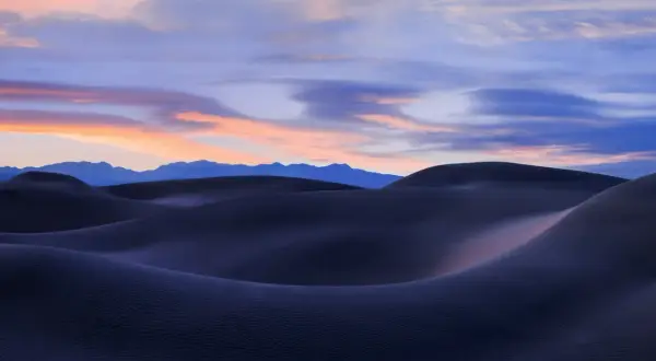Landscape photography sunset over dark sand dunes, with soft pink and blue clouds painting the sky above distant mountains.