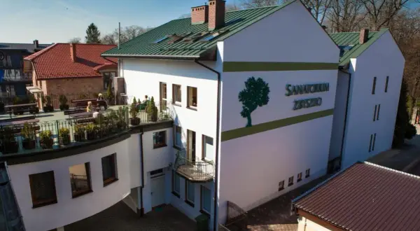 Sanatorium Zbysko w Busku-Zdroju, biały budynek z zielonym dachem i logo drzewa. Na tarasie wypoczywają kuracjusze.