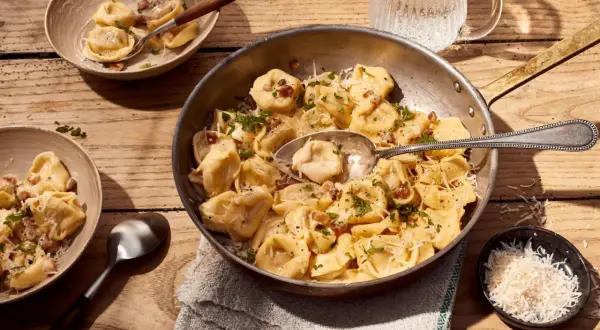 Herzhafte Tortellini Carbonara, garniert mit Petersilie und Parmesan, serviert in einer Pfanne und Schalen auf Holztisch.
