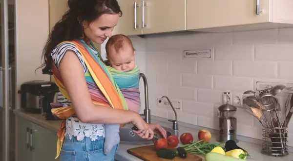Mamma förbereder mat vid amning i köket, med en bebis i bärsjal. Grönsaker ligger på bänken.