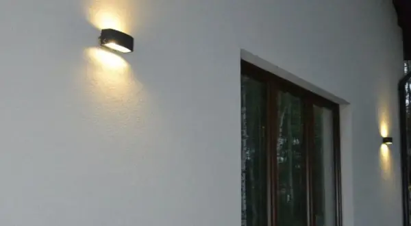 Nierówności na elewacji pod światło, tworzące ciekawe cienie. Dwa kinkietowe punkty świetlne oświetlają białą ścianę domu.