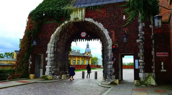 Kamienny łuk porośnięty bluszczem, przez który widać dziedziniec Wawelu. Dwie kobiety spacerują po brukowanym dziedzińcu.