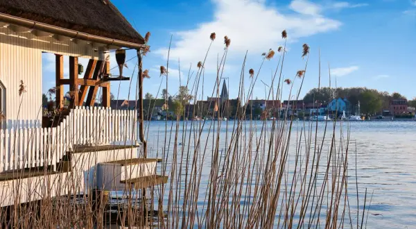 Reetgedecktes Haus am Plauer See mit Blick auf die Stadt und den Kirchturm. Schilf wiegt sich im Wasser.