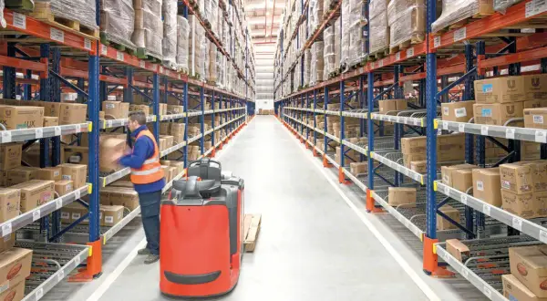 Pracownik magazynu, order picker, przenosi paczkę. Czy ciężko? Wysokie regały pełne towarów.