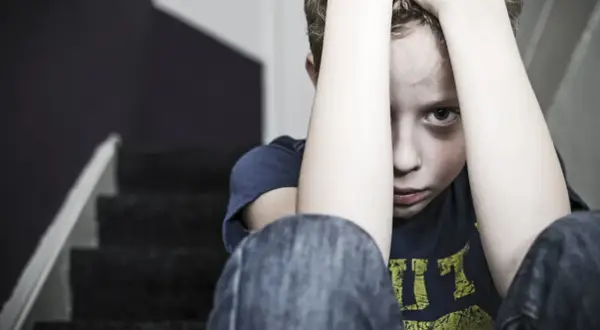 En pojke sitter på en trappa med händerna över huvudet, blicken tom. Bilden symboliserar känslan av obehandlad PTSD.